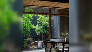 Un coin repas avec une table et des chaises, un petit vase avec des fleurs et une vue sur un jardin arboré, agrémenté d’une clôture en bois.