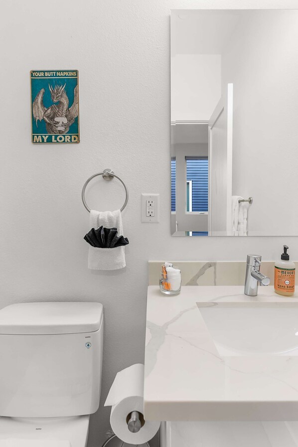 Un baño moderno con inodoro, lavabo y espejo. Una toalla cuelga de un perchero y hay un cartel en la pared.