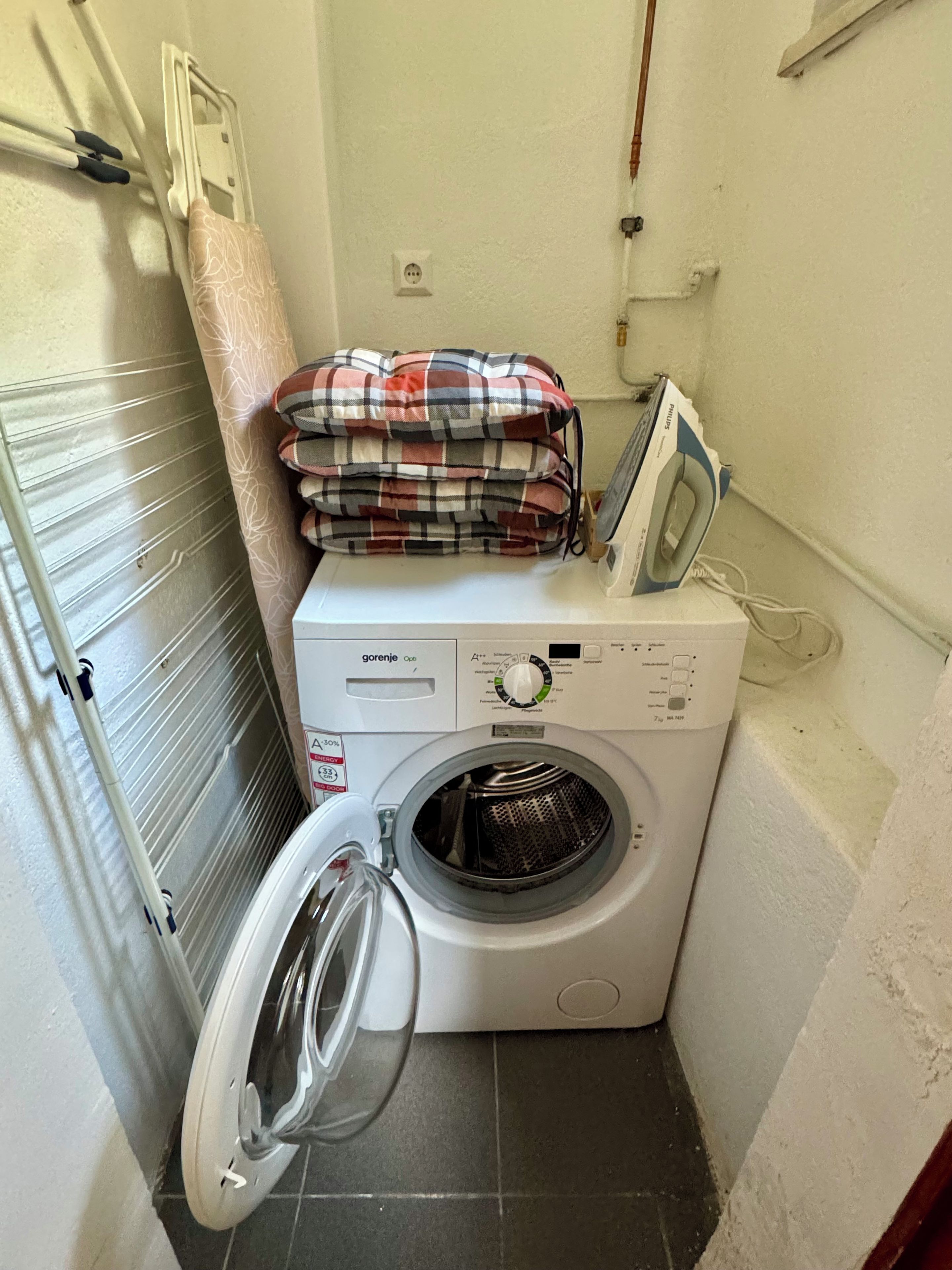 Eine kompakte Waschmaschine mit Bedienfeld, eine zusammengefaltete Decke und ein Bügeleisen darauf.