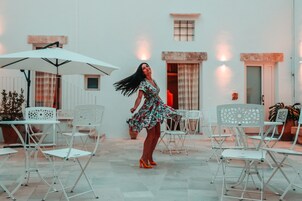 Una mujer con vestido floral y zapatos de tacón naranja bailando en un patio con sillas y mesas blancas, un paraguas blanco y un edificio con fachada azul claro al fondo.