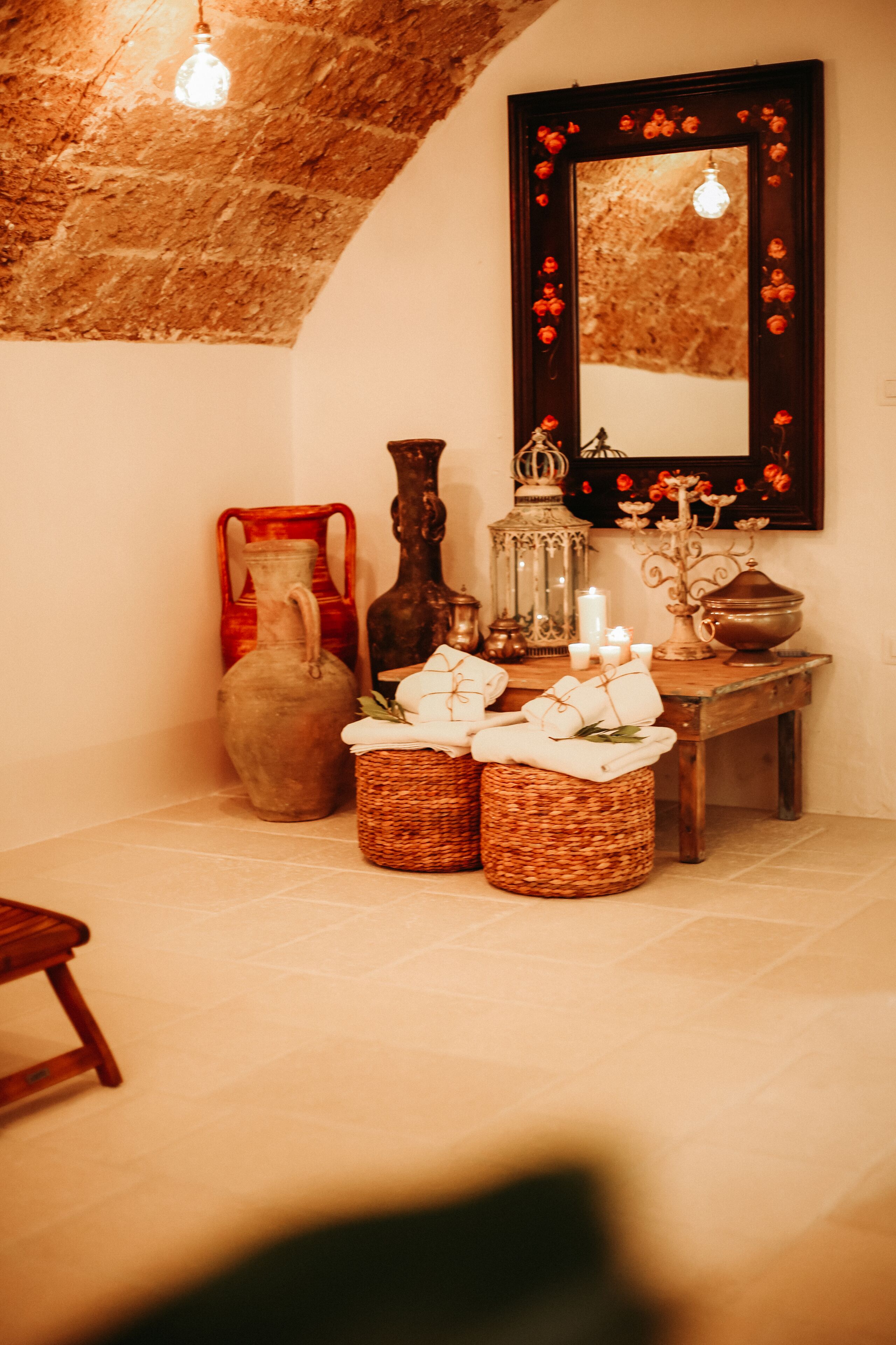 A rustic room with a stone ceiling, a wooden bench, a mirror with red flowers, and various decorative items on a table.