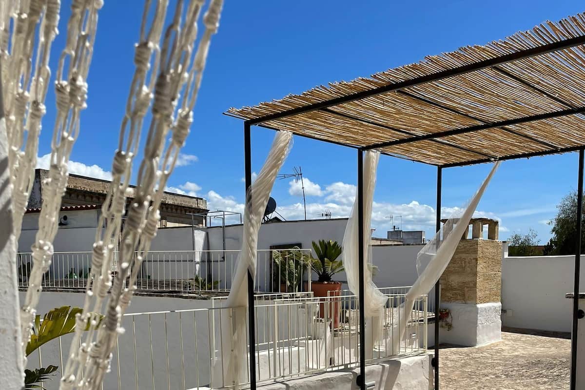 A rooftop patio with two wooden lounge chairs, a pergola with a thatched roof, and a view of neighboring buildings.