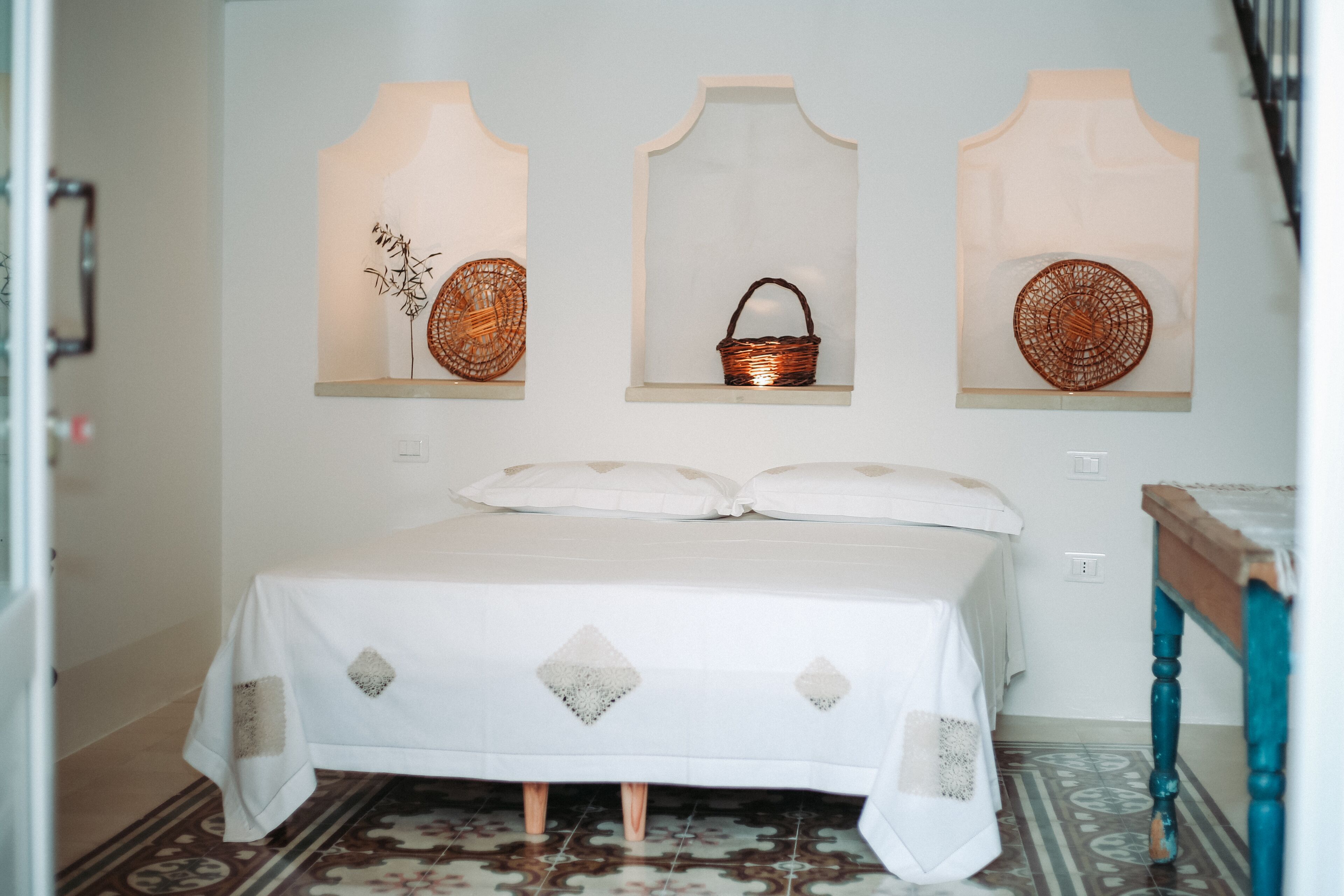 A bed with a white bedspread and pillows, a wooden nightstand, and three wall-mounted shelves with woven baskets and a woven bowl.