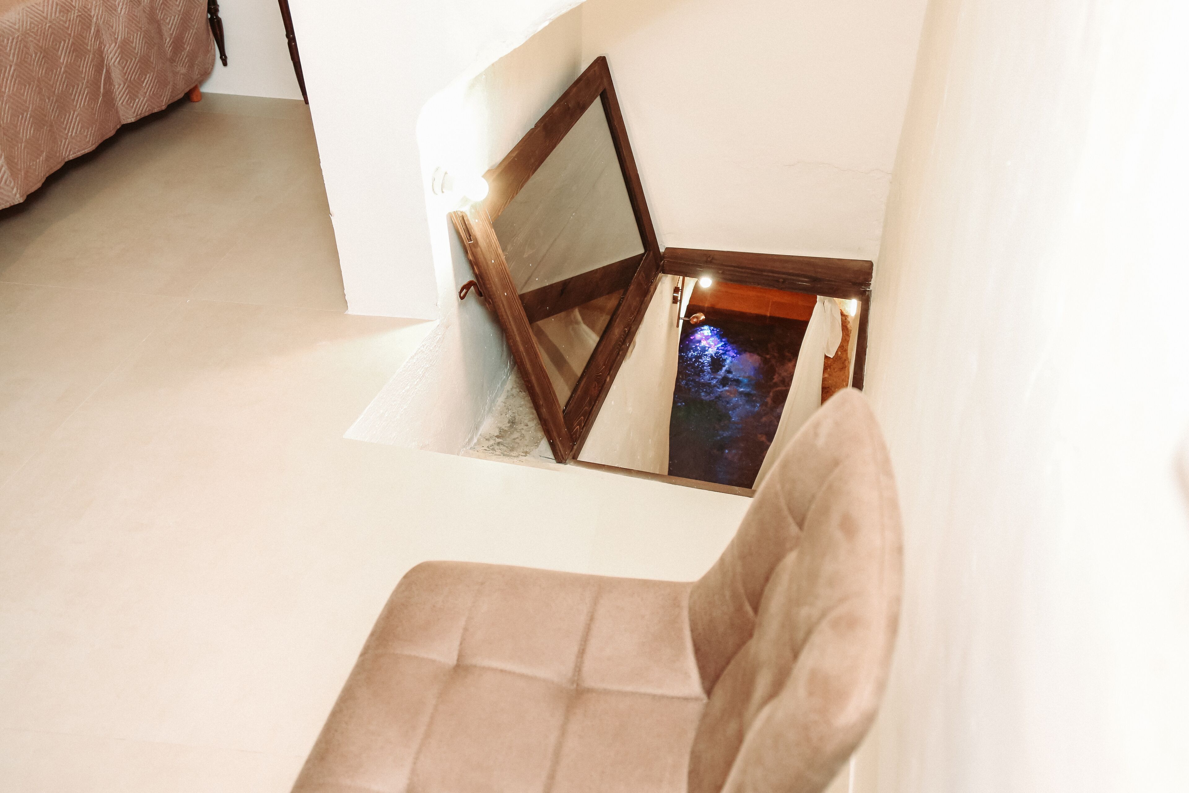 A corner of a room with a beige chair, a wooden-framed mirror, and a small window with a view of a night sky.