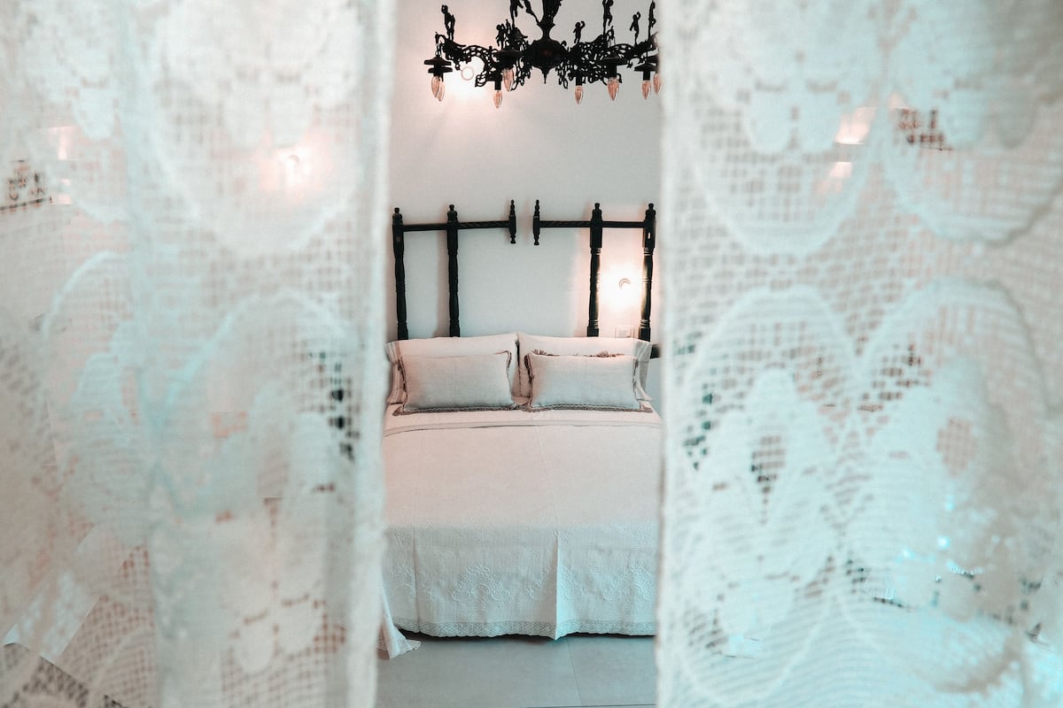 A bedroom with a bed, a chandelier, and lace curtains.