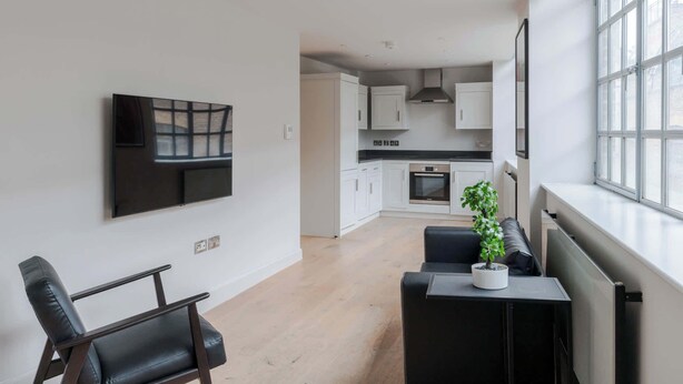 A modern kitchen with white cabinets, a black countertop, and an oven. There is a dining area with a black table and chairs, and a large window providing natural light. - Farringdon 1-Bed Apartment · Lift · Sleeps 3 (London)