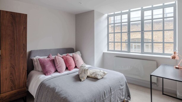Une chambre à coucher comprenant un lit, une armoire en bois, un radiateur et une fenêtre donnant sur des immeubles en briques.