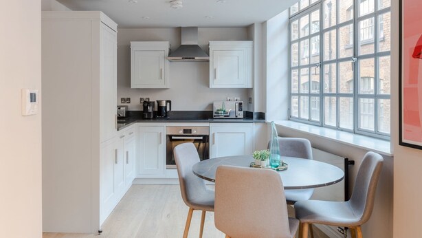 A modern kitchen with white cabinets, a black countertop, and a window with a view of buildings. There is a dining table with chairs and a small plant on it. - Clerkenwell 2-bed Apartment · Lift · Sleeps 4 (London)