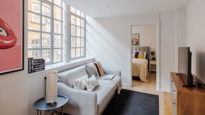 A modern living room with a grey sofa, a small side table, and a large window with a view of buildings. - Clerkenwell 2-bed Apartment · Lift · Sleeps 4 (London)