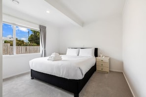 A bedroom with a bed, a nightstand, a window with curtains, and a view of the outside. - Spacious Renovated 3BR in Ellerslie I Zodiak Stays (Auckland)