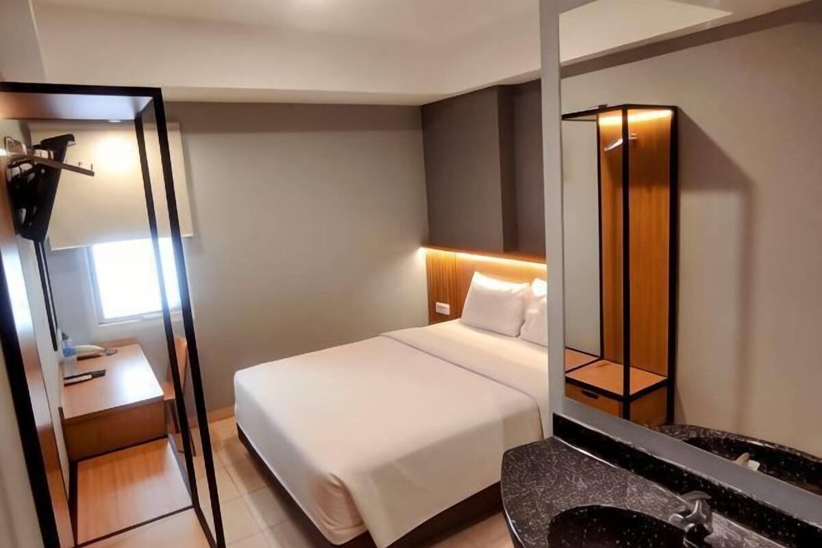 A hotel room with a bed, a desk, a mirror, and a bathroom with a sink and mirror.