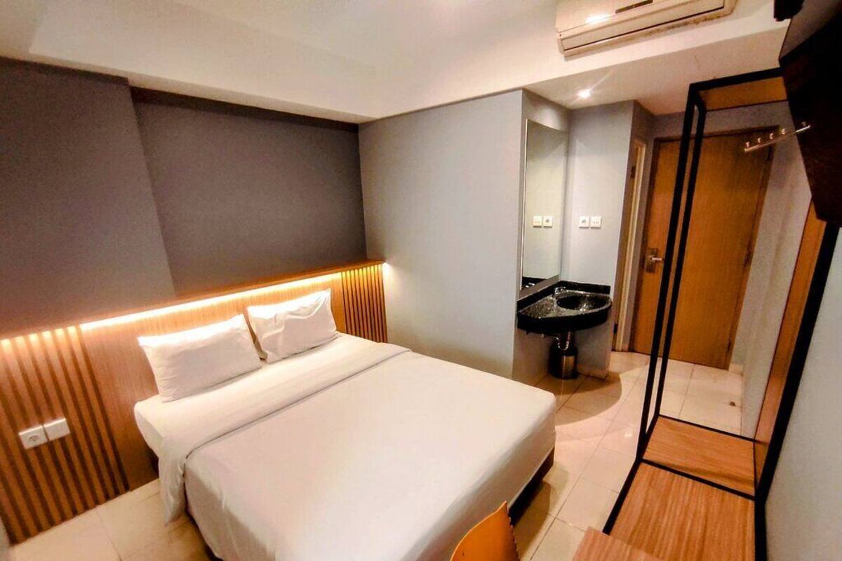 A modern hotel room with a large bed, a small table, a mirror, and a bathroom with a sink and toilet.