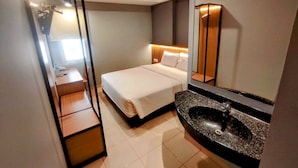 Una habitación de hotel con cama, baño con lavamanos y espejo, y una estantería de madera.