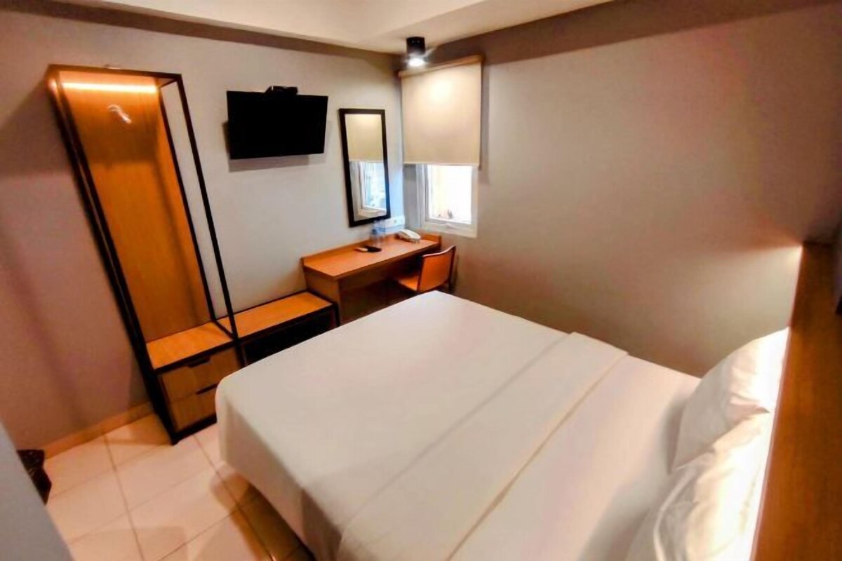 A hotel room with a bed, a desk, a chair, a TV mounted on the wall, and a window with blinds.