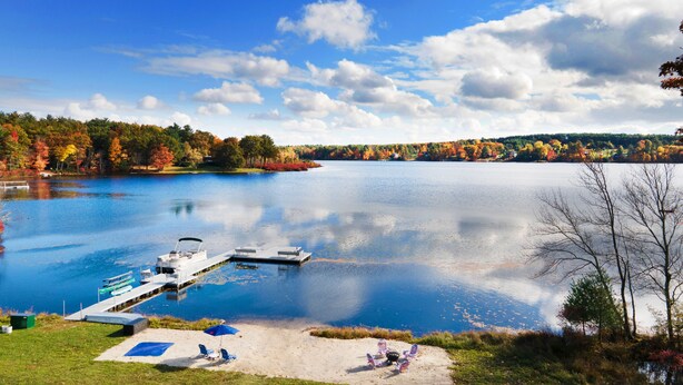 A lakeside view with a dock, a boat, and a sandy area with flags. - SwimSpa | Sport Courts | Lake | Fishing | Firepit (Highland Lake)