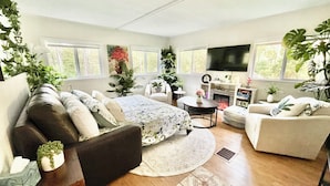 A spacious living room with a large bed, two sofas, a coffee table, and a fireplace. There are multiple windows providing natural light and a TV mounted on the wall. - SwimSpa | Sport Courts | Lake | Fishing | Firepit (Highland Lake)