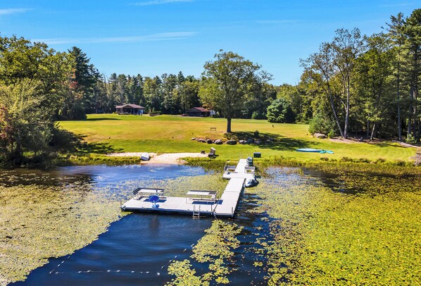 A dock extending into a pond surrounded by a grassy area with trees and a building in the background.