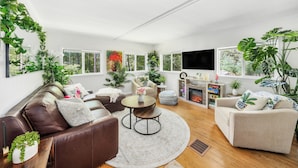 A modern living room with a brown leather sofa, beige armchairs, a round coffee table, and a fireplace. There are multiple potted plants and a large window providing a view of greenery outside. - SwimSpa | Sport Courts | Lake | Fishing | Firepit (Highland Lake)