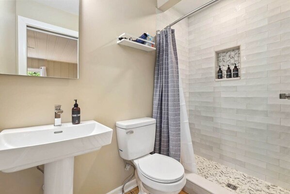 Un baño con lavamanos, inodoro y cortina de ducha blancos. Hay artículos de aseo en el lavamanos y un estante sobre el inodoro.