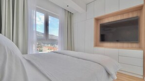 Een moderne slaapkamer met een groot bed, een televisie bevestigd aan een houten paneel, en uitzicht op de stad door het raam.