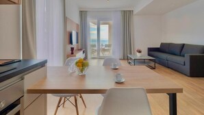 Een moderne hotelkamer met een houten eettafel, witte stoelen, een grijze bank en uitzicht op zee door grote ramen.