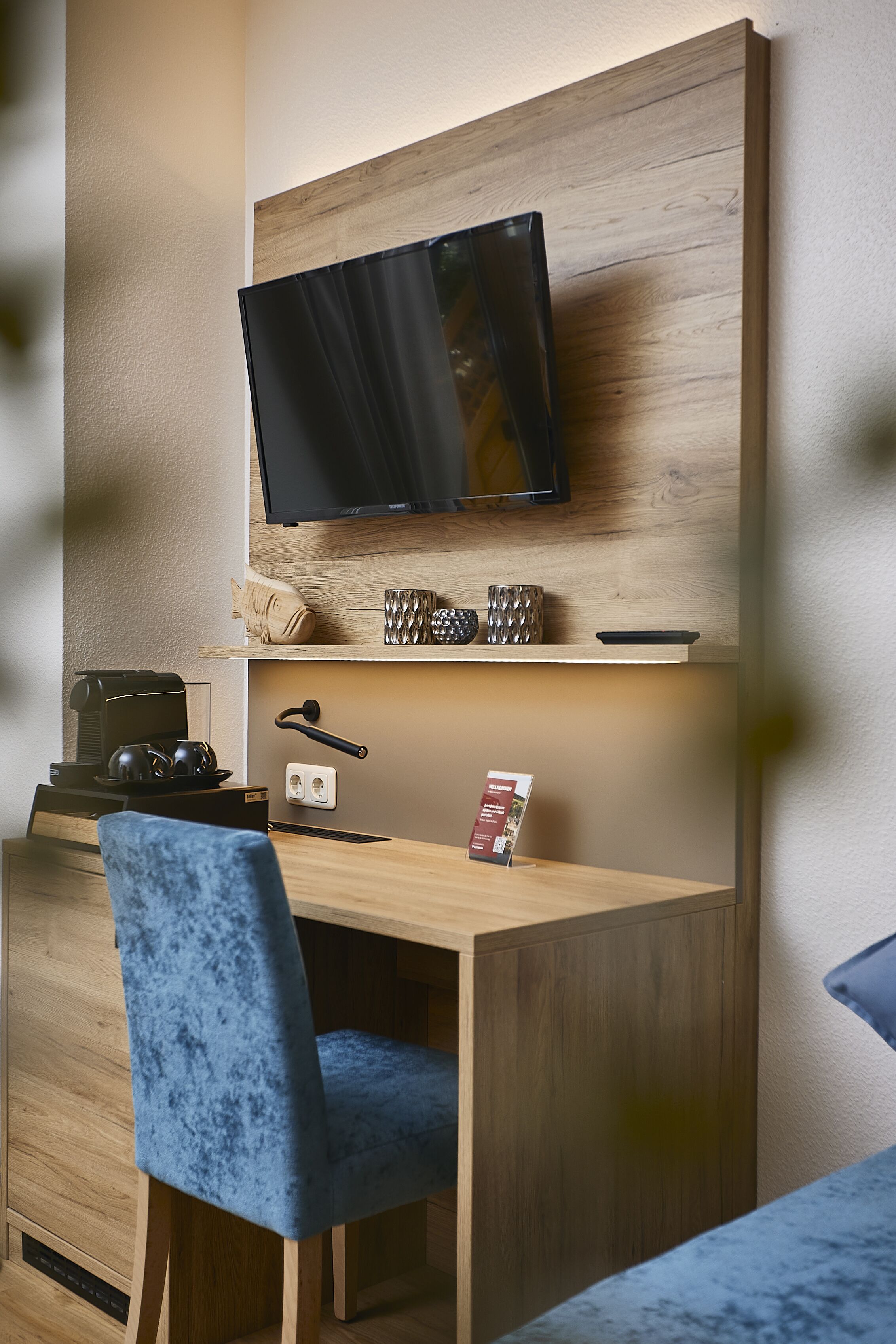 Habitación de hotel moderna con un escritorio de madera, una silla azul, un televisor de pantalla plana montado en la pared y una cafetera.