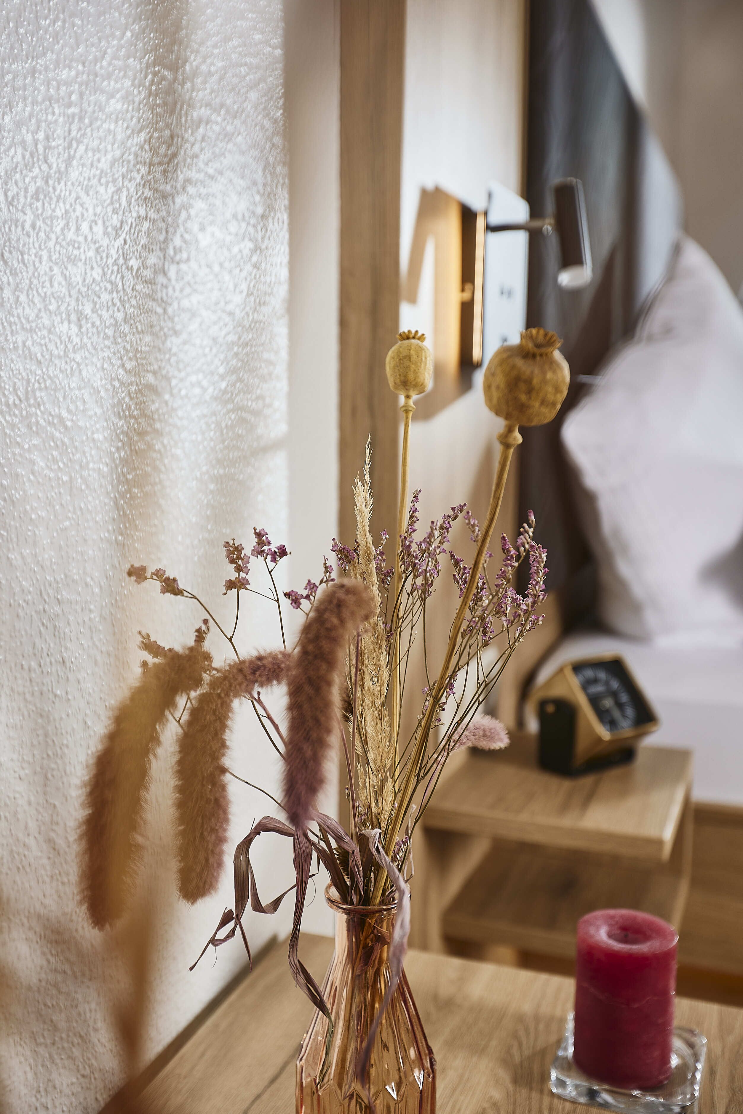 A vase with dried plants on a wooden table, a red candle, and a clock in the background.