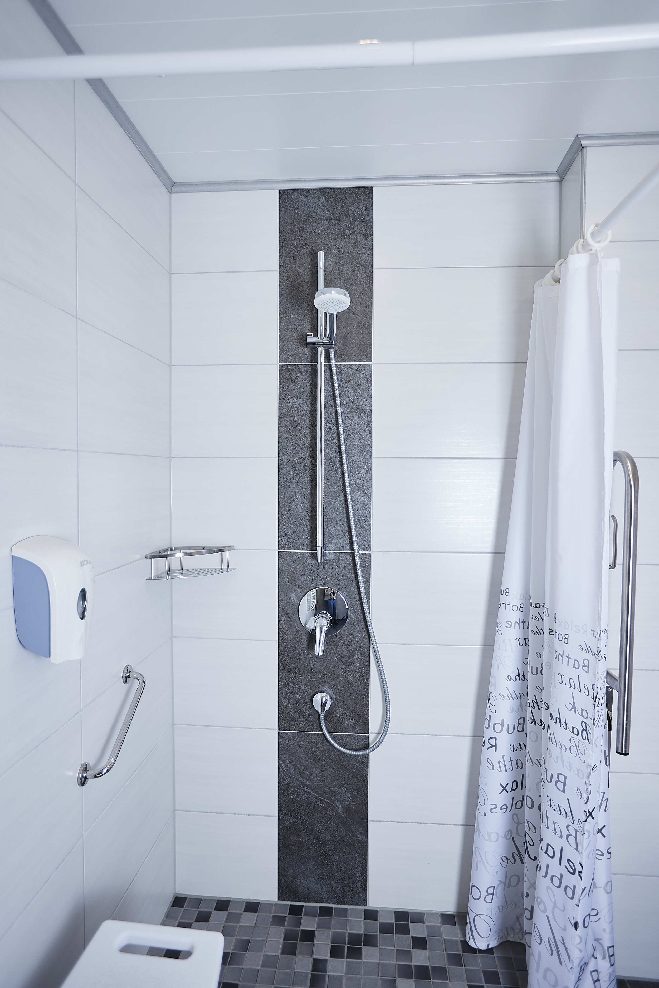 Een moderne badkamer met witte tegels, een douche met een handdouche, een toilet met een leuning en een handdoekdispenser.