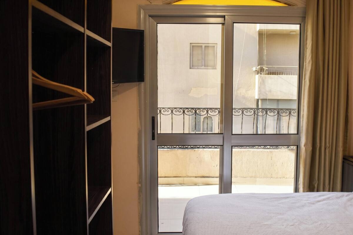 A room with a bed, a wardrobe, a television mounted on the wall, and a balcony with a railing.