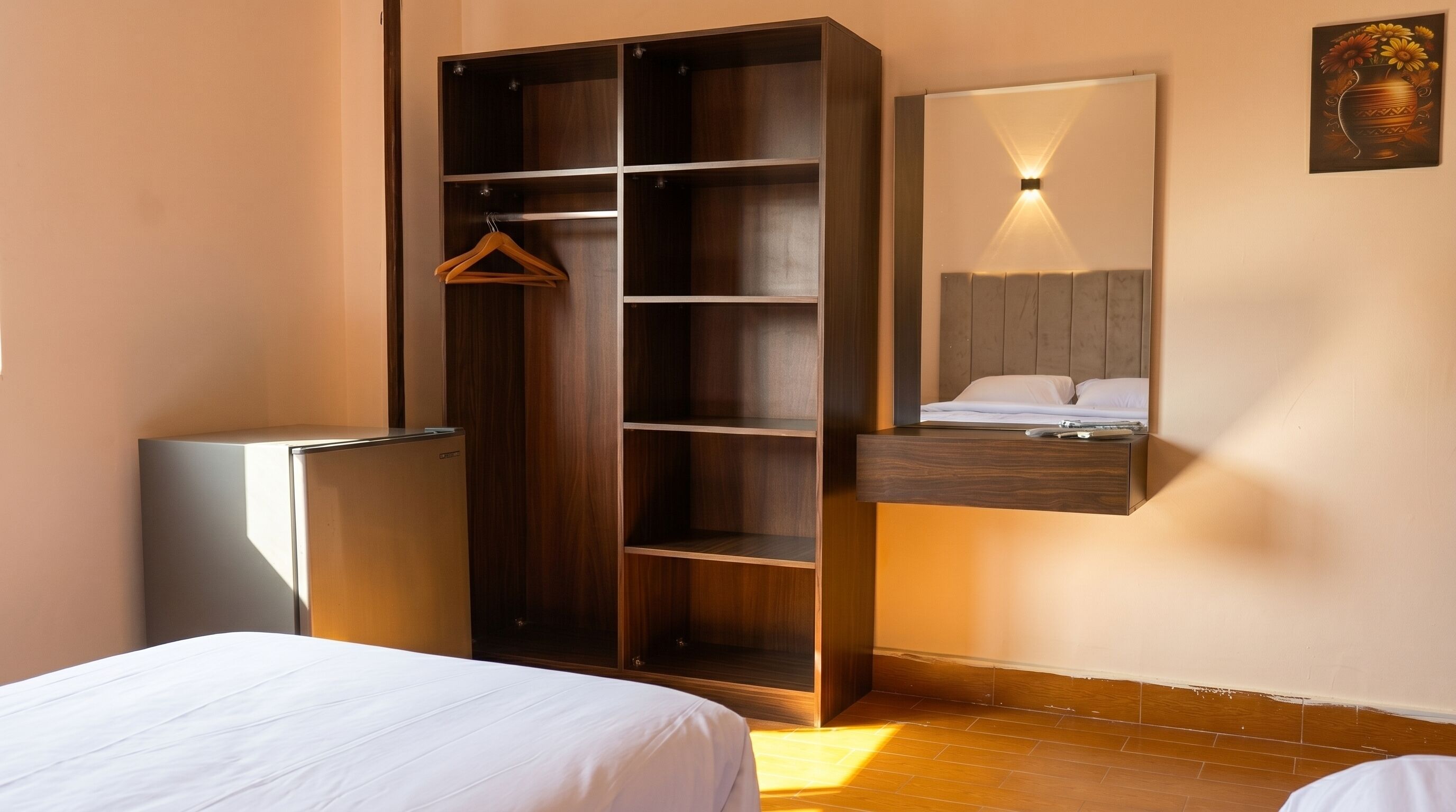 A hotel room with a wardrobe, a bed, a small fridge, and a mirror with a wall-mounted light.