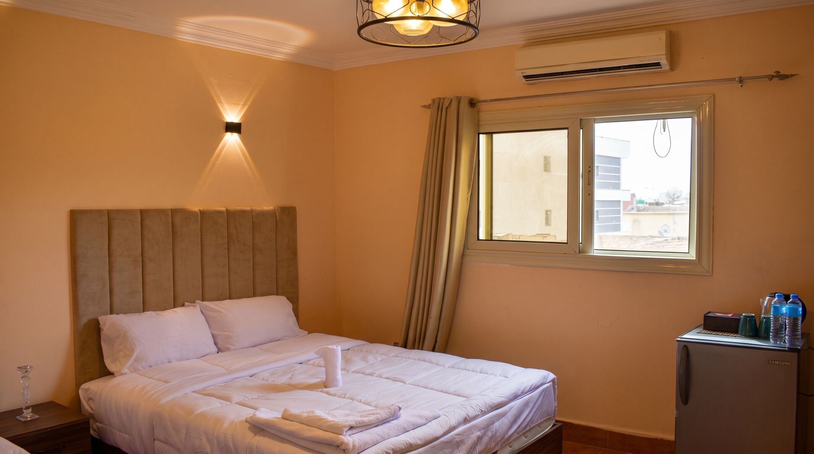 Une chambre d’hôtel comprenant un lit, une table de chevet, un bureau avec une bouteille d’eau, un climatiseur et une fenêtre avec des rideaux.