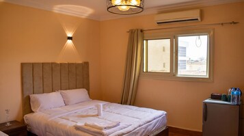 Habitación de hotel con una cama, una mesita de noche, un escritorio con una botella de agua, aire acondicionado y una ventana con cortinas.