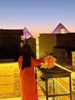 A person in a long orange garment standing in front of a metal grill with food items.
