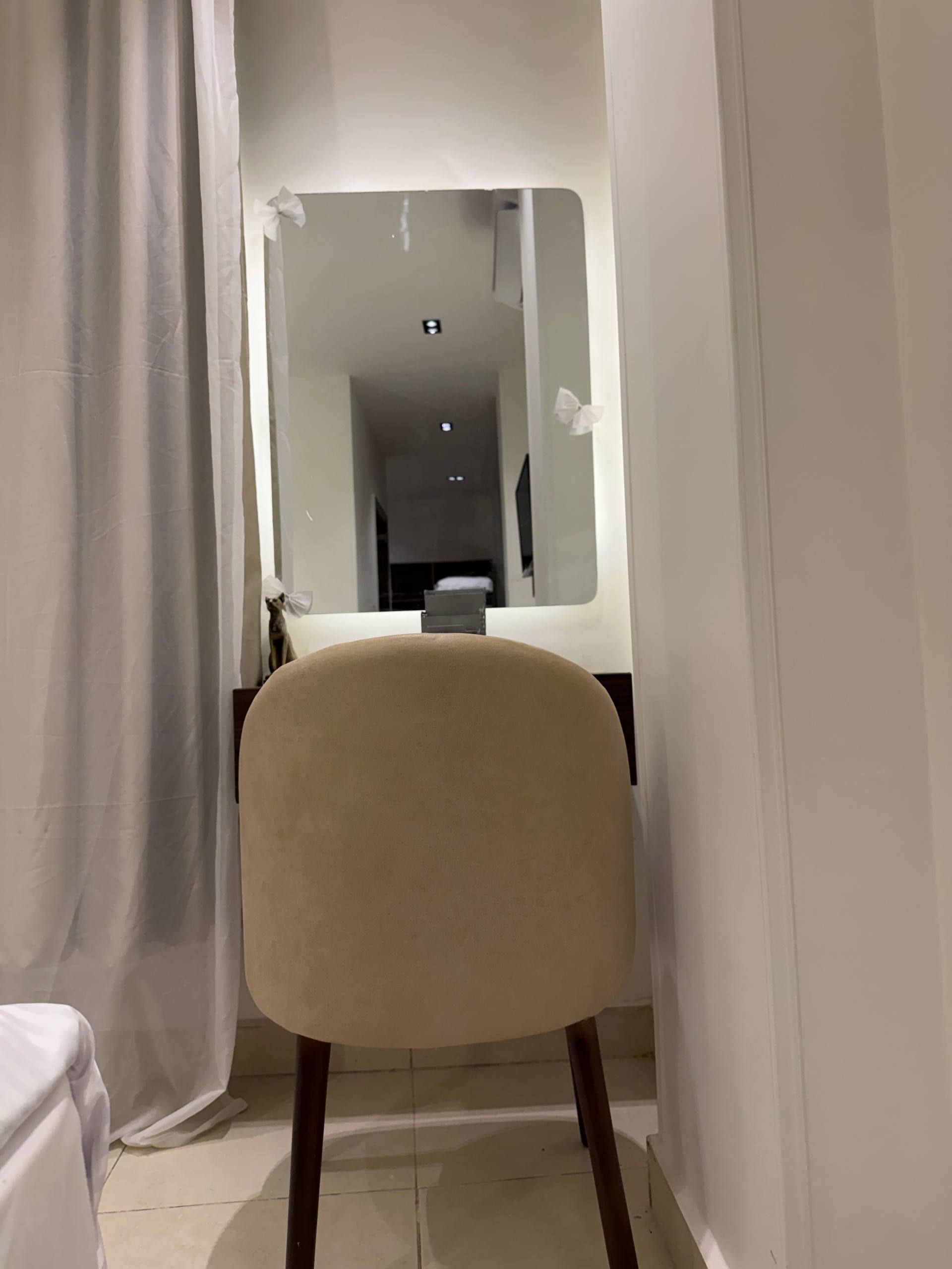 A narrow hallway with a chair and a mirror reflecting a room with a bed and a TV.