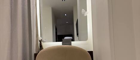 A narrow hallway with a chair and a mirror reflecting a room with a bed and a TV.