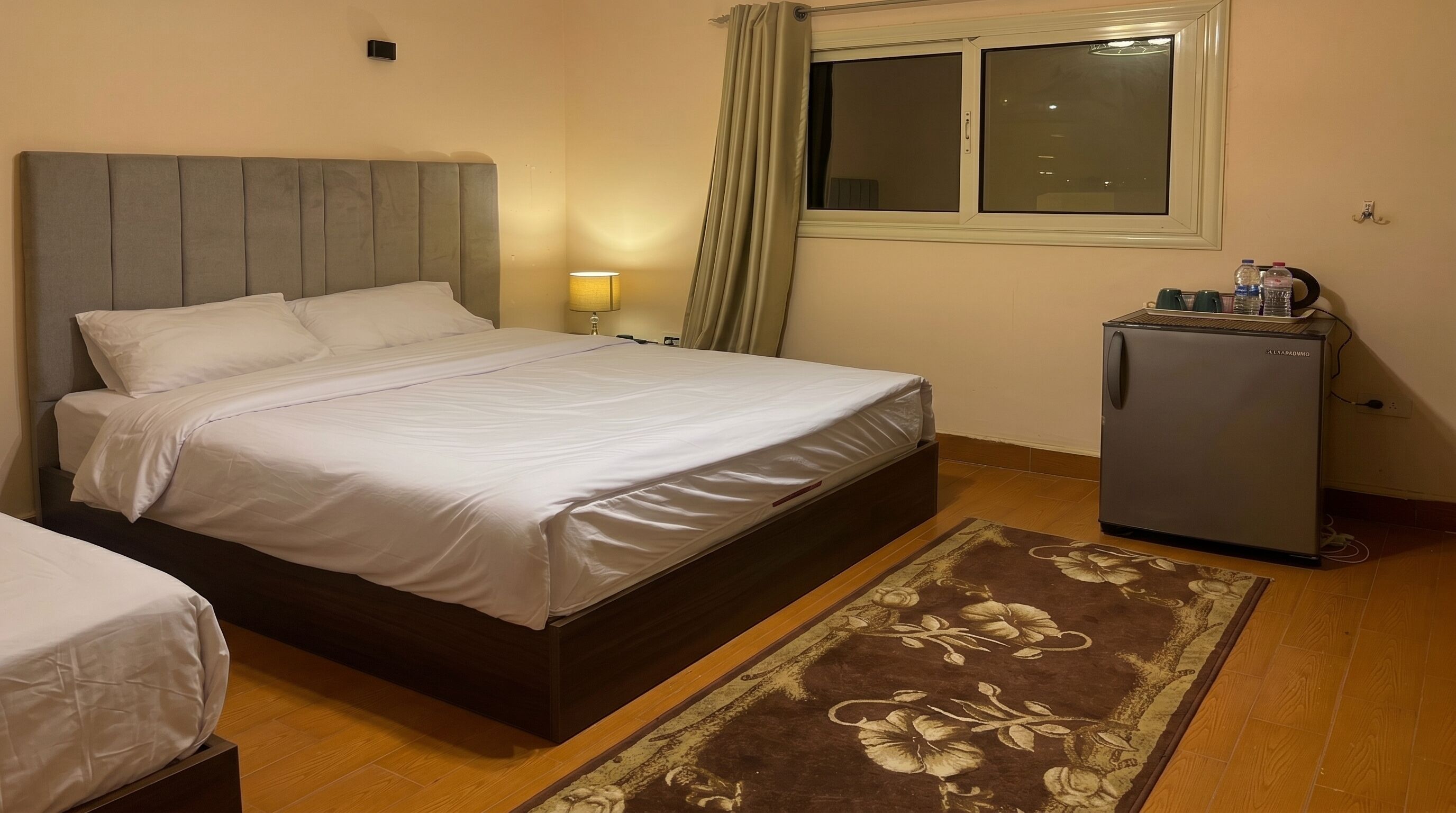 Un dormitorio con una cama, una mesita de noche, una lámpara, una ventana con cortinas, un refrigerador y una alfombra estampada.
