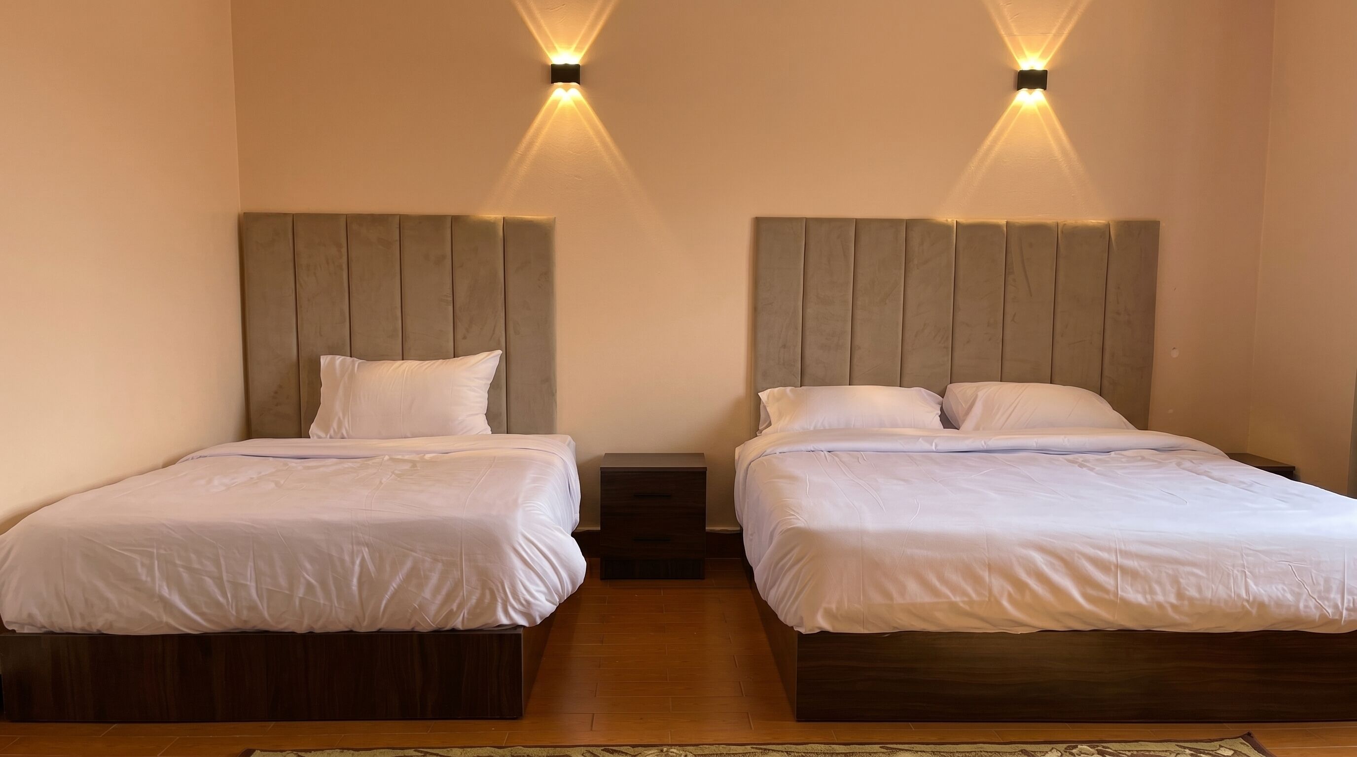 A room with two beds, each with a wooden headboard, a nightstand, and a wall-mounted light fixture.