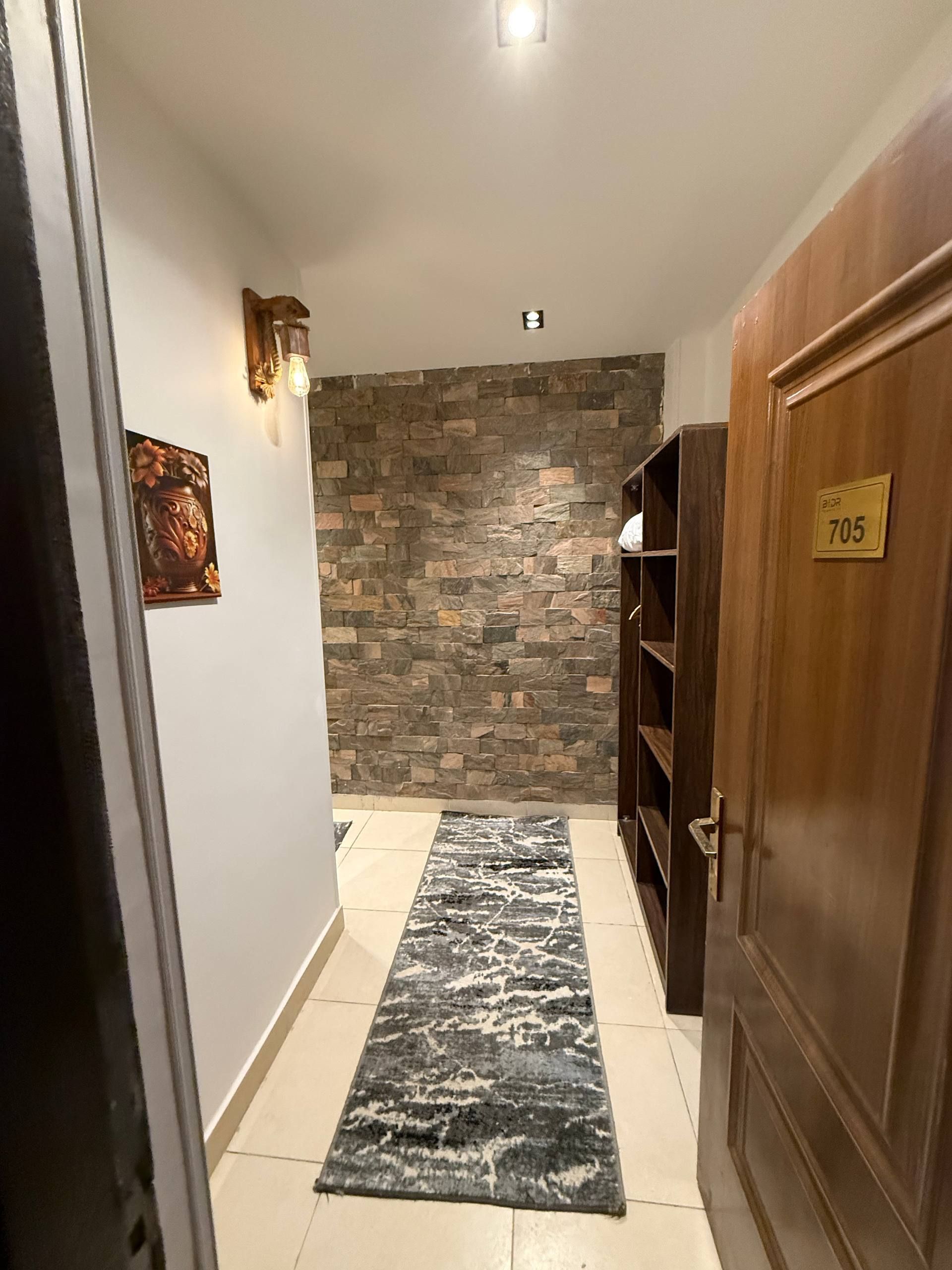 A hallway with a stone wall, a wooden door with a number plate, and a patterned rug.