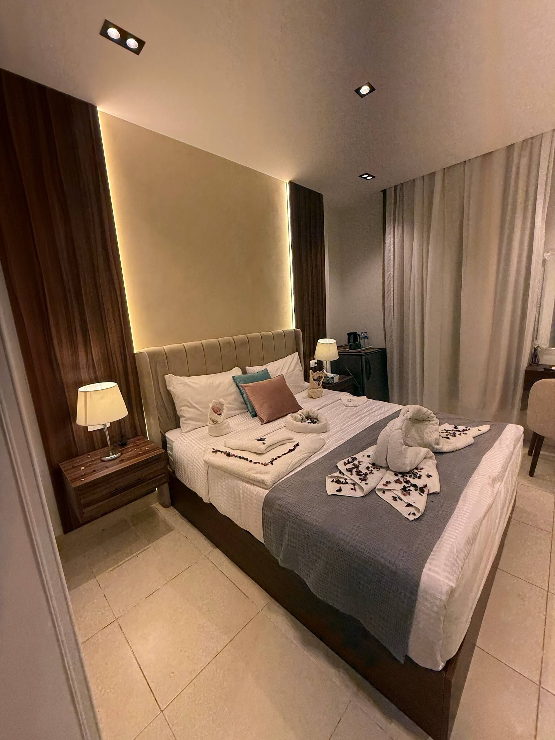 Habitación de hotel con una cama grande, lámparas de noche, una mesita de noche y vistas de la ciudad a través de cortinas.