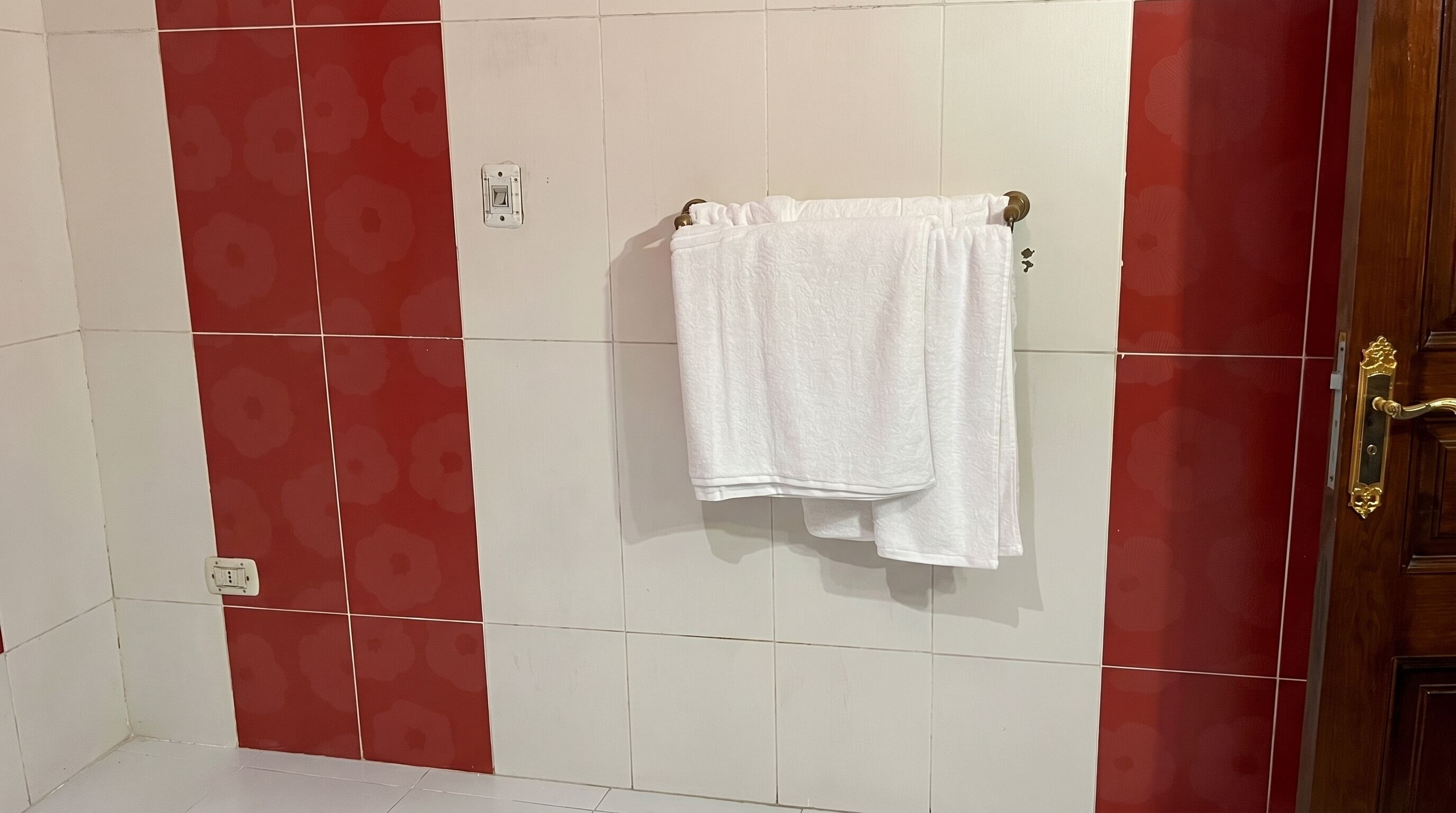 Une salle de bain carrelée de rouge et de blanc, un porte-serviettes avec deux serviettes et une porte dotée d’une poignée en laiton.