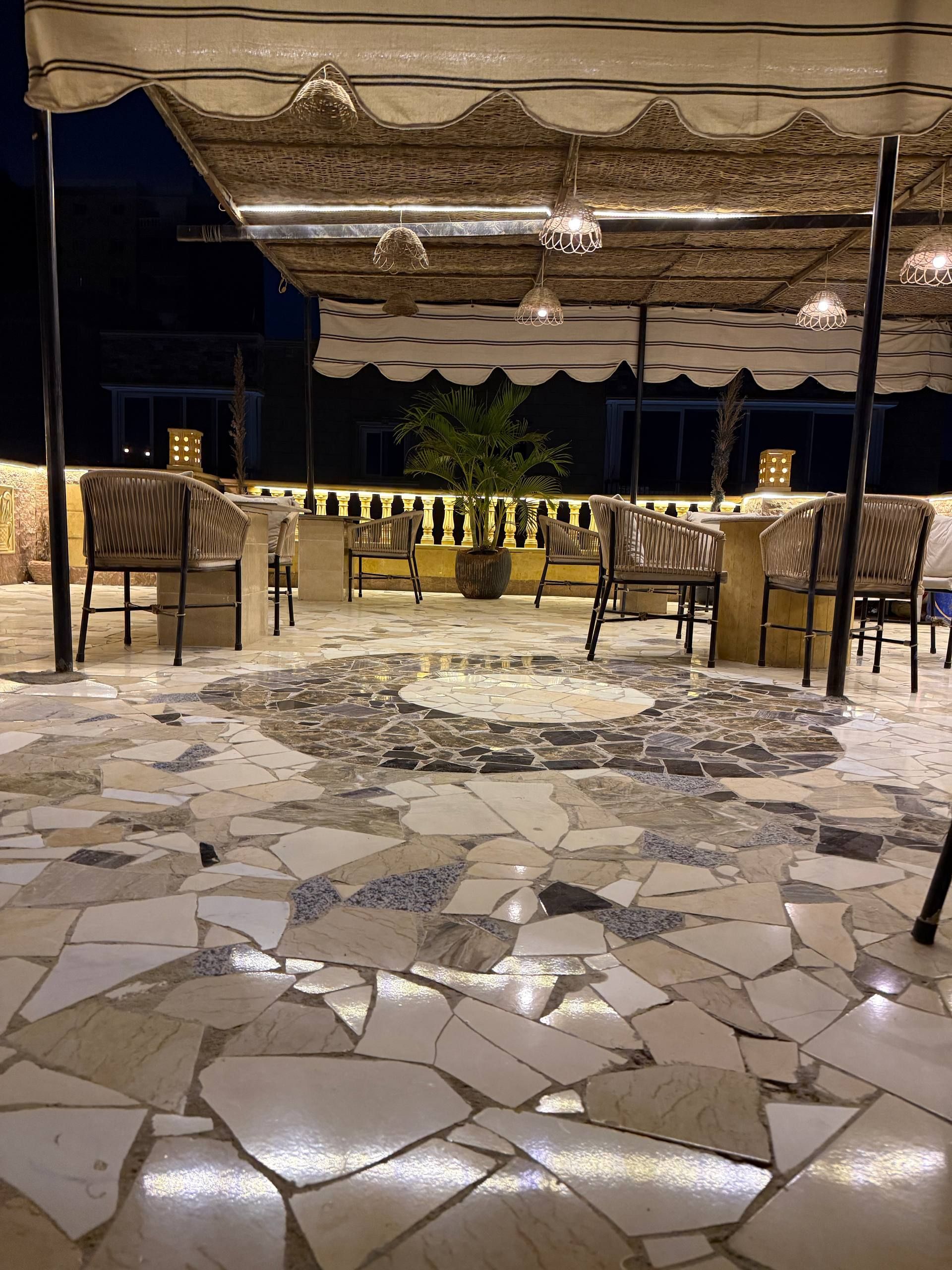 Un espacio de estar al aire libre con suelo de mosaico, sillas de mimbre, una planta central y un toldo.