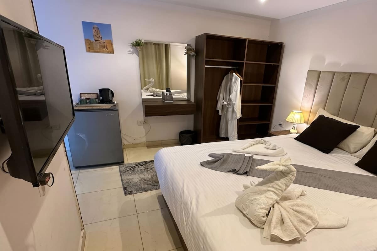 A hotel room with a bed, a wardrobe, a desk with a lamp, and a small refrigerator.