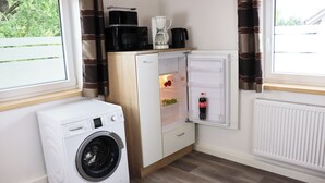 Une cuisine compacte équipée d’un lave-linge, d’un réfrigérateur avec compartiment congélateur, d’une machine à café et d’une bouilloire posées sur le plan de travail.