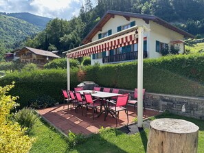 En uteplats med ett bord och stolar, en pergola och utsikt över ett berg och några hus.