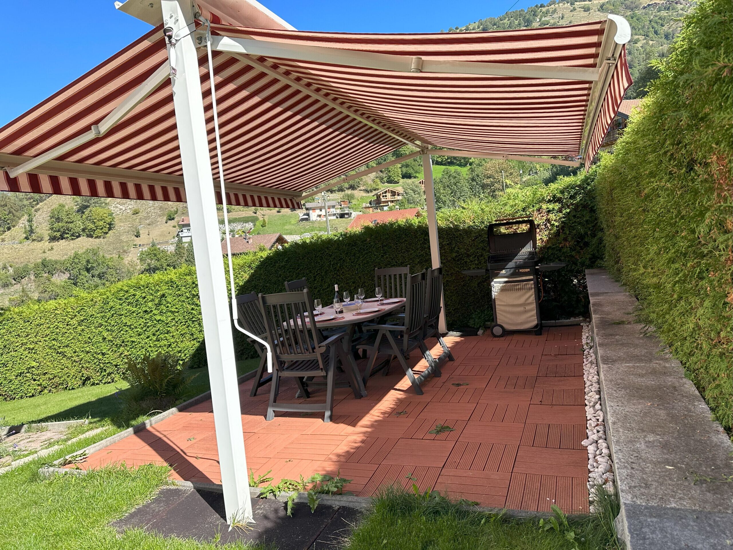 A patio with a table and chairs under a striped awning, a grill, and a view of a hillside.