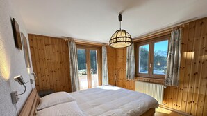 A bedroom with a wooden floor, a bed with white bedding, a hanging light fixture, and large windows with curtains.