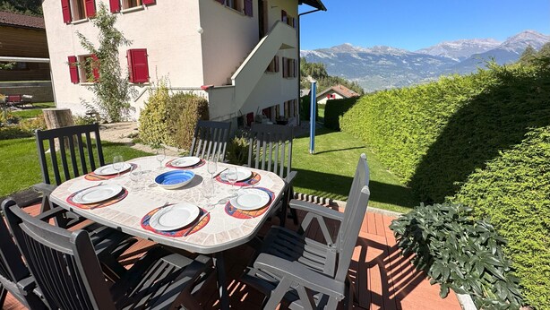 A dining table set outdoors with a mountain view, a staircase leading to a second floor, and a well-maintained garden.