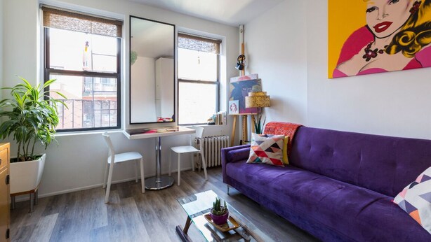 A modern living room with a purple sofa, a glass coffee table, a white desk, and a large window with a view of buildings.