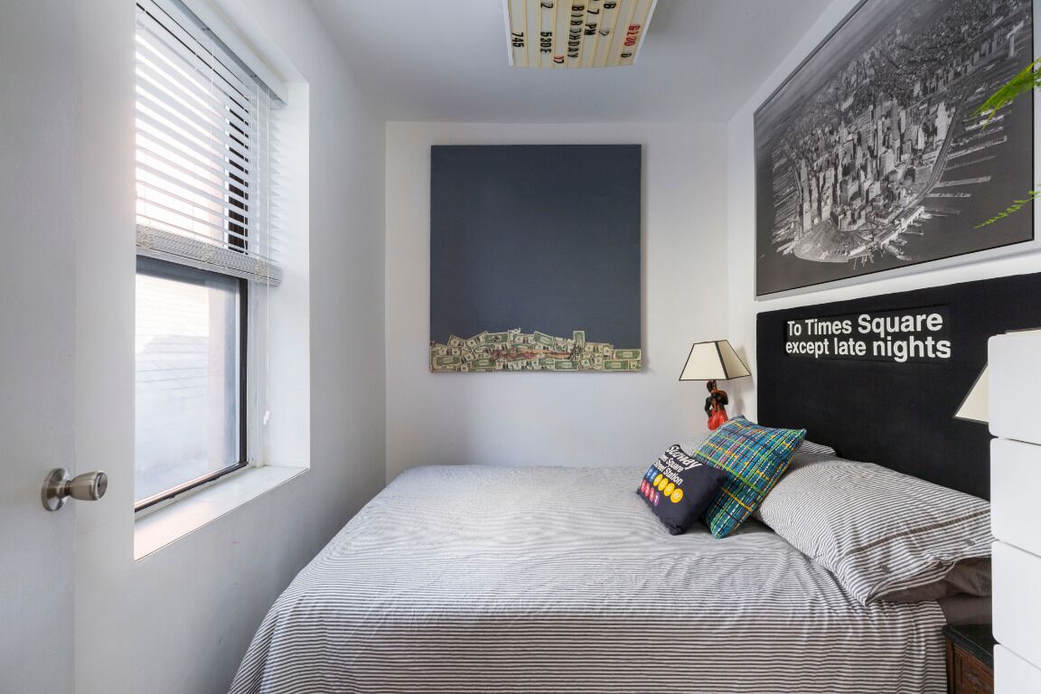 A bedroom with a bed, a window with blinds, a cityscape painting, and a poster on the wall.