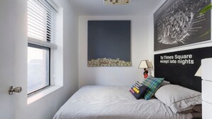 A bedroom with a bed, a window with blinds, a cityscape painting, and a poster on the wall.